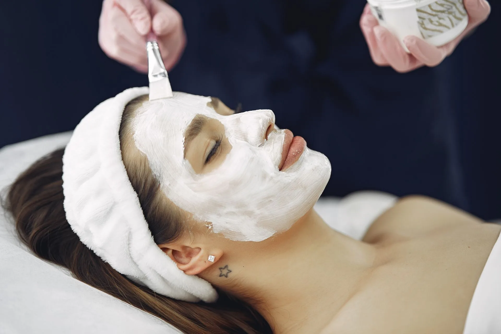 Client relaxing with eyes closed while a facial mask is applied with a brush